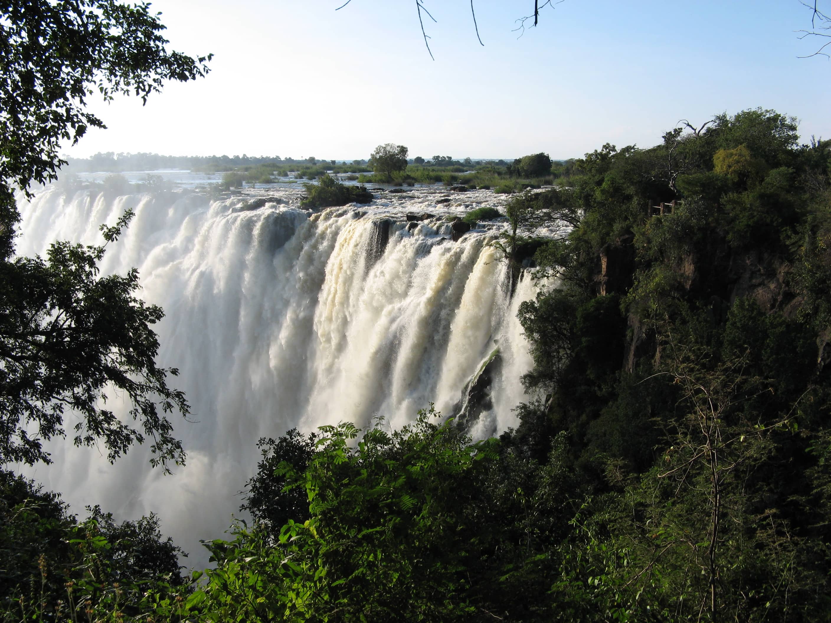 Victoria Falls thundering over the gorge in bright sunshine, framed by lush green trees in Zambia.jpg