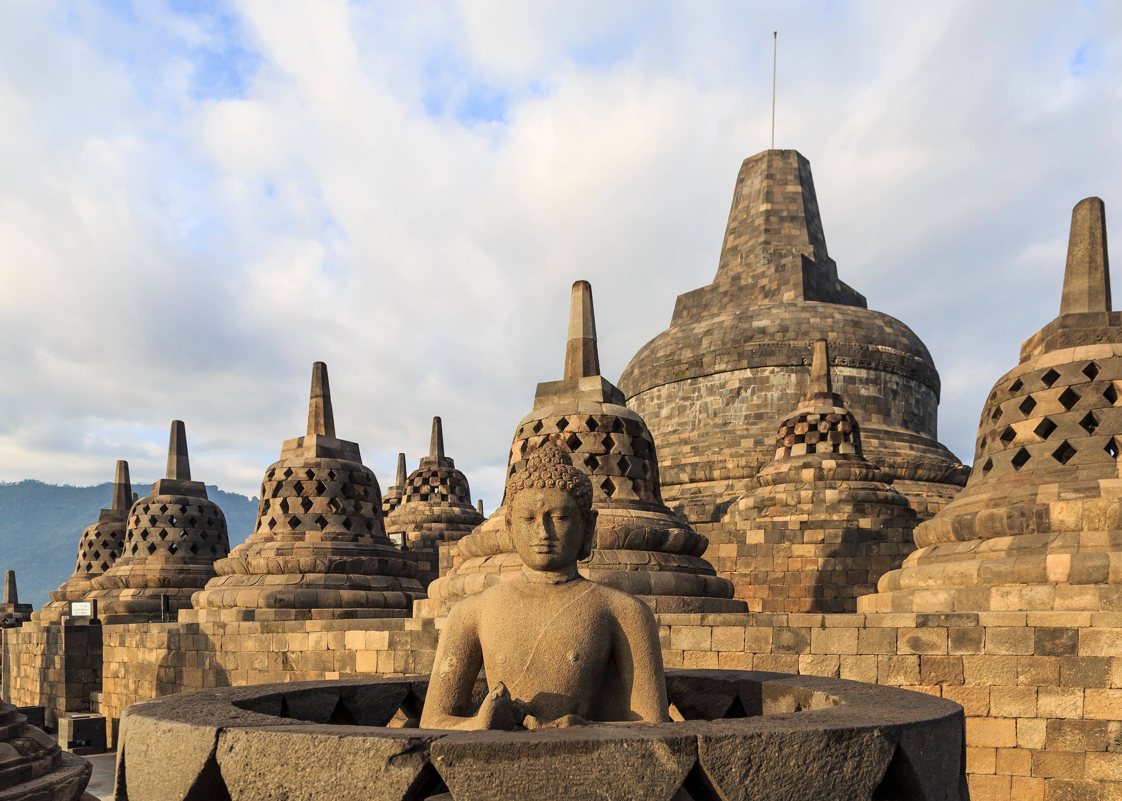 Sunlit stone stupas and a Buddha statue on the upper terrace of Borobudur temple, Java, Indonesia